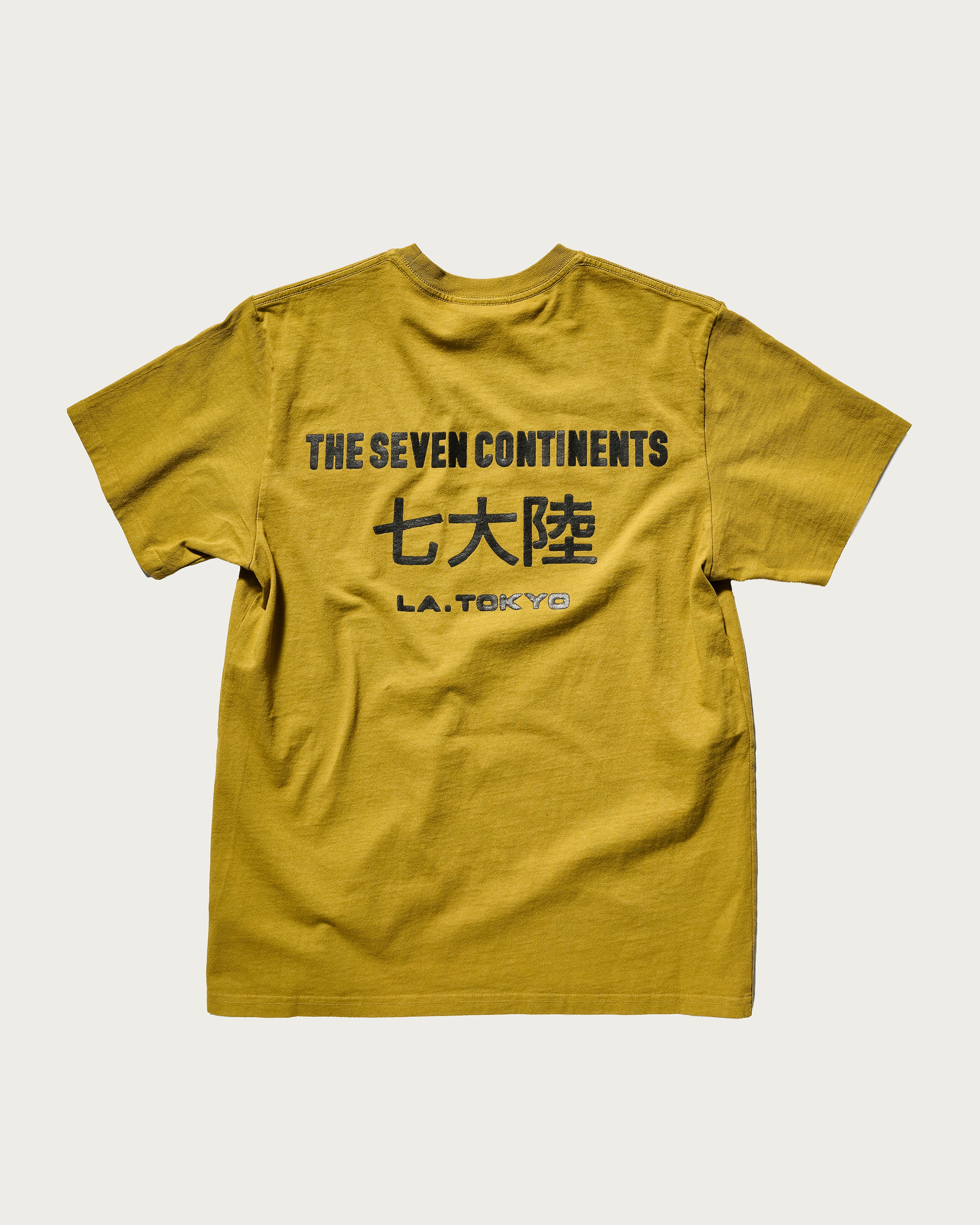 The T7C The Seven Continents Timo Tee in Mustard features a puff screen printed logo and black text on the back reading THE SEVEN CONTINENTS with Japanese characters and L.A. TOKYO underneath. Shirt is shown laid flat on a plain background.