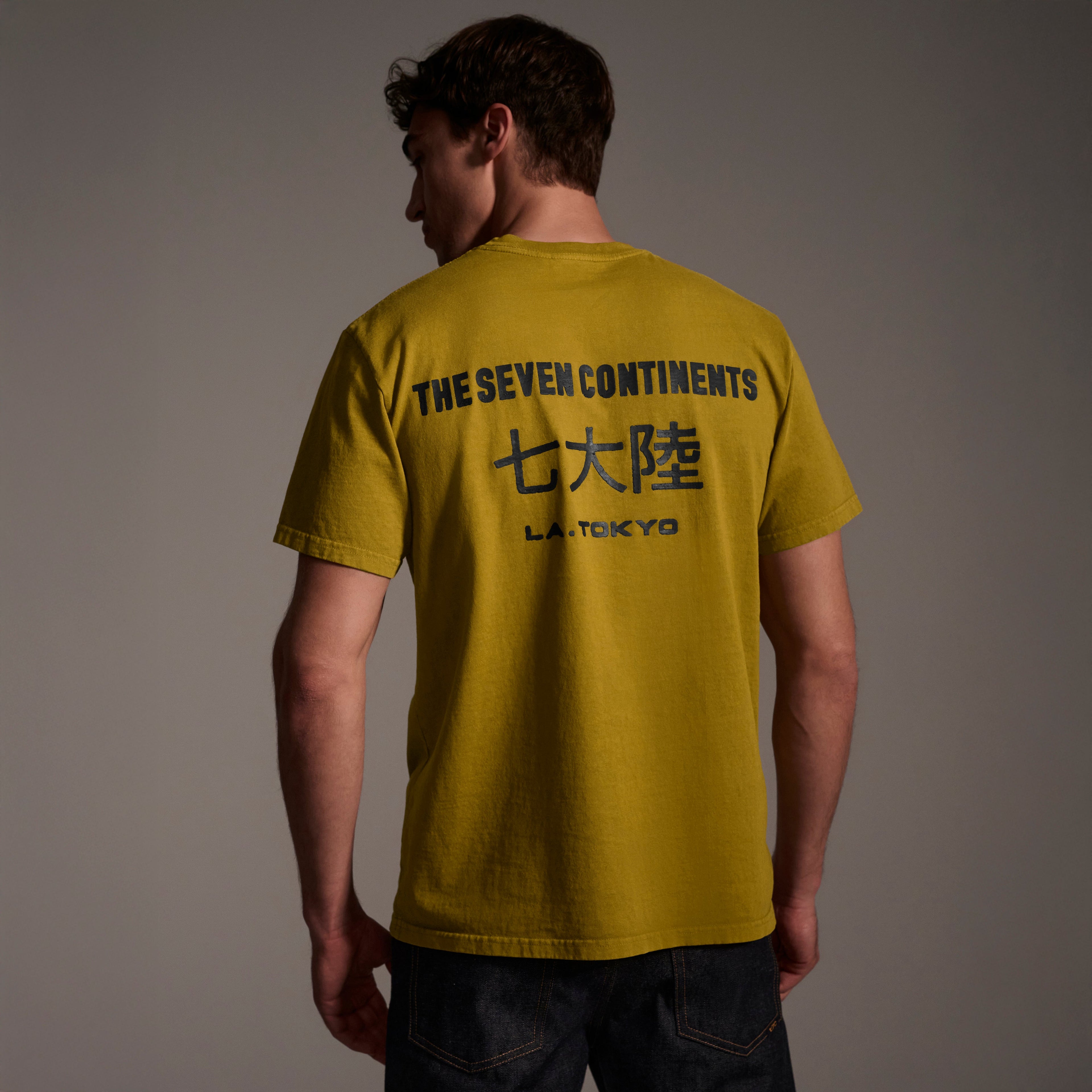 A man faces away, wearing The Timo Tee in Mustard by T7C The Seven Continents. The puff print on the back features THE SEVEN CONTINENTS, Japanese characters, and L.A. TOKYO. He pairs it with dark jeans against a neutral background.