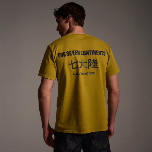 A man faces away, wearing The Timo Tee in Mustard by T7C The Seven Continents. The puff print on the back features THE SEVEN CONTINENTS, Japanese characters, and L.A. TOKYO. He pairs it with dark jeans against a neutral background.