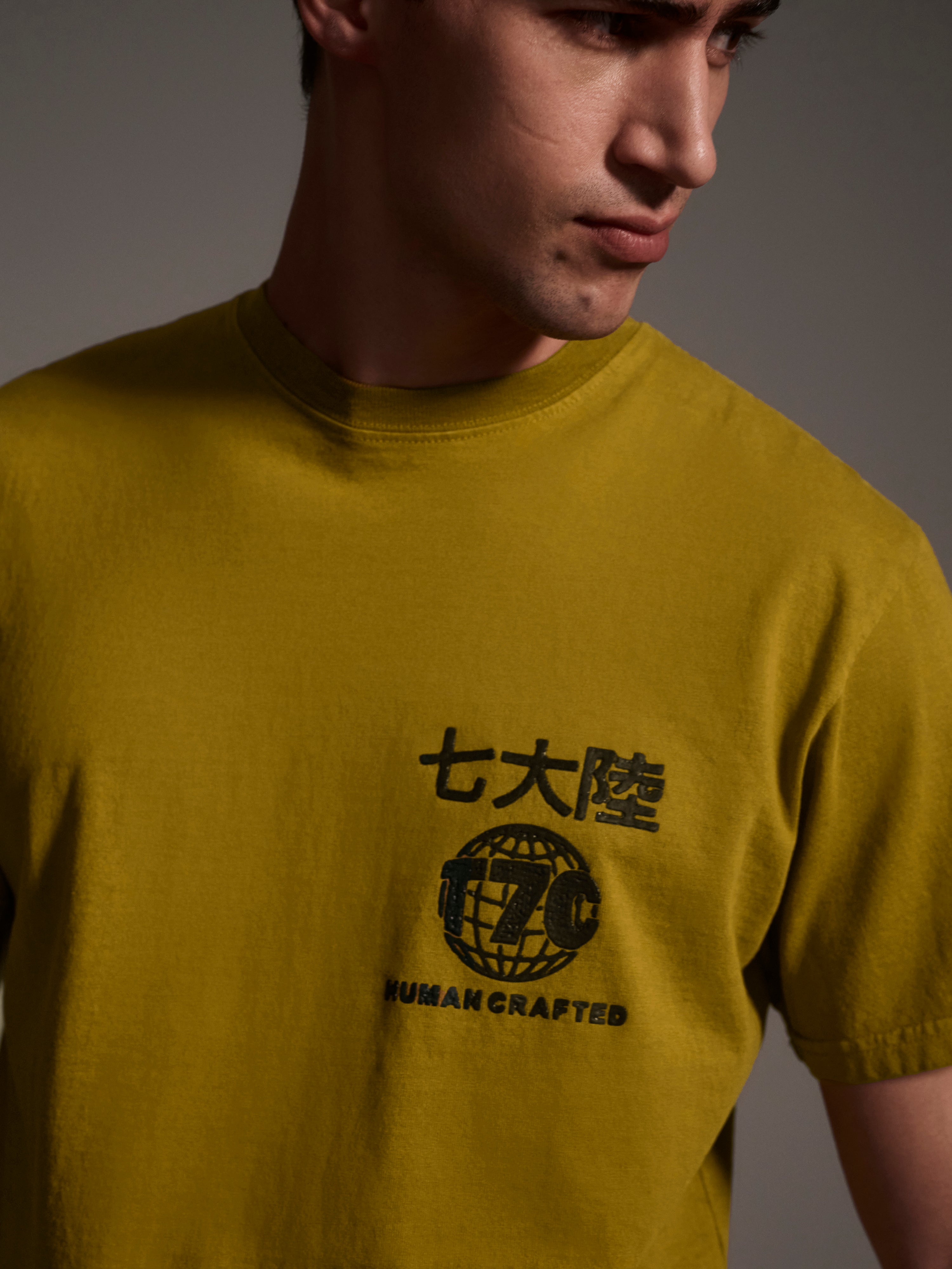 A detail image of a male model wearing a mustard t-shirt with a logo detail